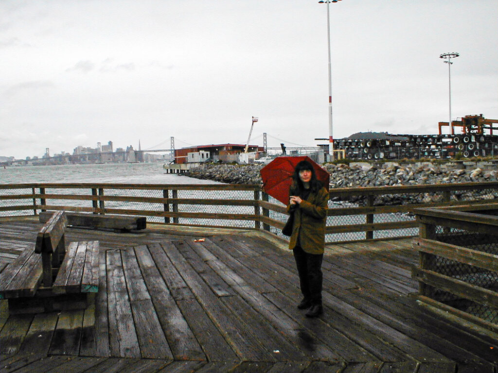 Overcast day, woman with umbrella on pier, SF skyline in distance.
Photo: T. Christensen.