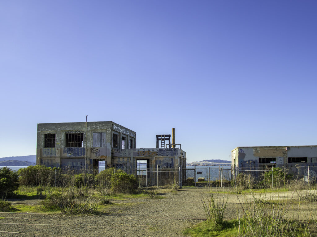 Abandoned buildings at Ferry Point with scrubby vegetation and the bay behind under deep blue sky.